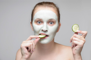 Girl with green cream on face. Beautiful young woman with homemade mask biting  a cucumber