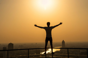 Fototapeta premium Silhouette of man standing with extend arms action over the cityscape background at sunset time with lens flare,Lifestyle and freedom concept