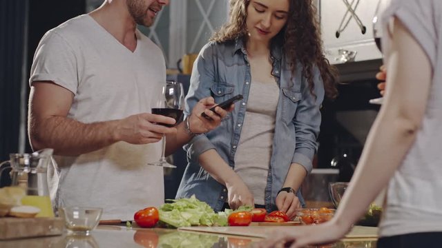 Tilt up shot of young man reading something funny on smart phone and holding glass of wine while having cooking party with friends in kitchen