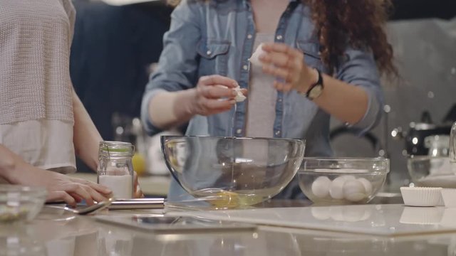 Tilt up of two young women preparing mixture for baking: one woman cracking egg and pouring it into glass bowl and another girl adding salt