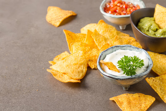 Snack for a party, chips with a tortilla, nachos with sauces: salsa with tomatoes, sour cream and guacamole. Mexican food. Dark background.  Copy space