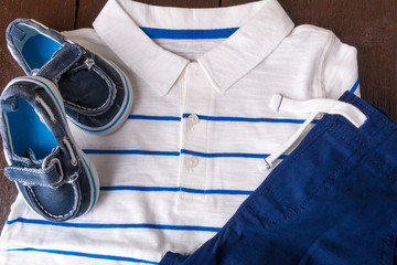 Blue boat shoes near shorts and white striped polo on brown wooden background. Boy outfit. Top view.