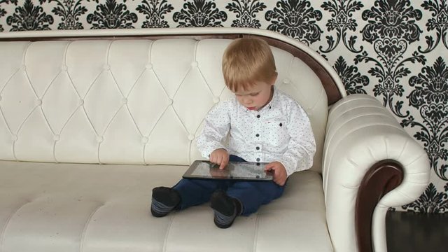 Little blonde boy playing with tablet sitting on a leather sofa, slow motion.