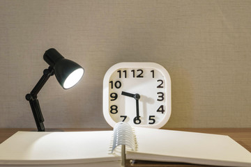 Closeup small lamp with light with blurred book and white alarm clock on wood desk textured background
