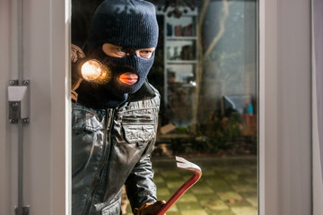 Masked burglar with flashlight and crowbar looking into glass window of house