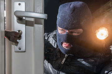 Masked burglar holding flashlight while trying to break window lock using crowbar at night