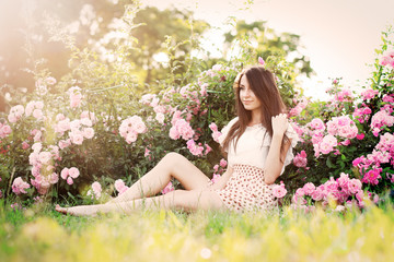 Obraz na płótnie Canvas красивая девушка в коротком платье сидит на траве рядом с розовыми розами в саду, теплая тонировка 