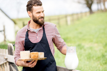 Portrait of a handsome farmer in apron standing with goat cheeses and milk outdoors on the green meadow background