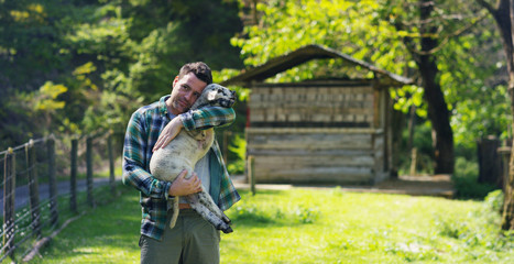 A young handsome and smiling farmer, holding a lamb young cub, has the experience to follow and care for live animals, on the background of nature and a barn, concept: ecology,livestock,bio,farming © Kitreel