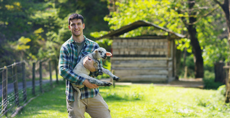 A young handsome and smiling farmer, holding a lamb young cub, has the experience to follow and...