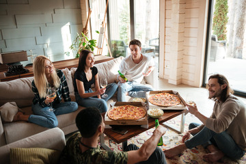 Cheerful young friends eating pizza and talking in living room