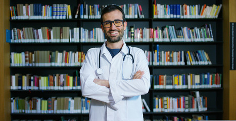 A young and beautiful doctor in a library smiling happy and holding books after doing a search and...