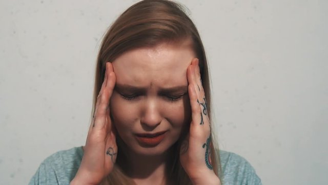 Suffering young caucasian girl in grey shirt with tattoos, ear flesh tunnels and birthmark on cheek have head ake in front of white wall, close up portrait