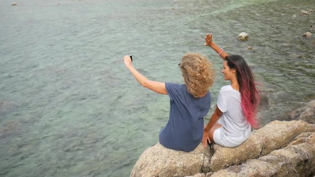 Two Young Carefree Hipster Girls Making Selfie Photo with Mobile Phone and Sitting on Rocks near Sea. HD Slow Motion Back View. Koh Phangan, Thailand.
