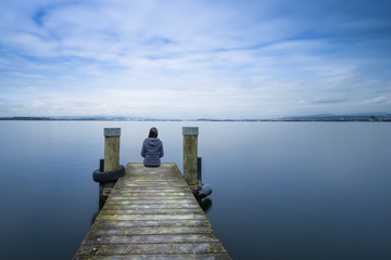 Overcast. Self reflection in magical world of fantasy. One woman sits on a wooden pier. Cloudy...