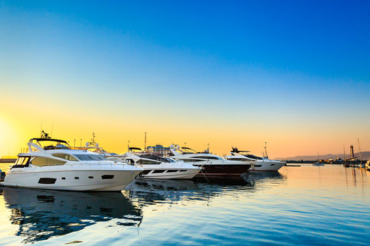 Fototapeta Luxury yachts docked in sea port at sunset. Marine parking of modern motor boats and blue water. Tranquility, relaxation and fashionable vacation.