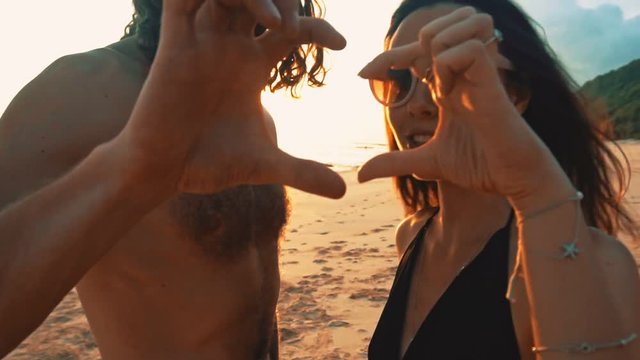 Happy young couple making heart shape with hands at the sunset on at sunset on a lonely beach