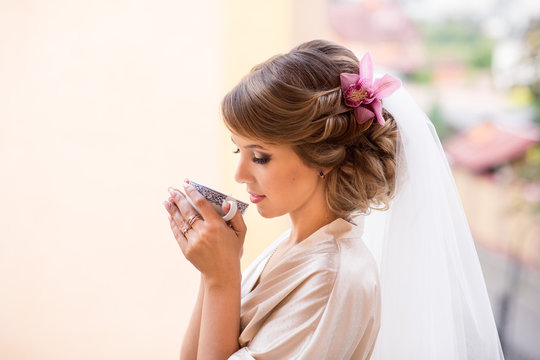 Beautiful Bride With Cup Of Coffee At Wedding Morning. Beauty Bridal Makeup And Hairstyle With Orchid Flower