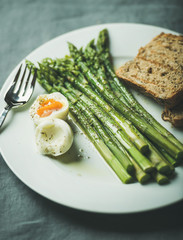 Cooked asparagus with soft-boiled egg, grilled bread and herbs on white round plate over grey textile background, selective focus. Clean eating, weight loss food concept