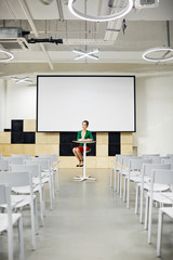 Young woman sitting by table in lecture-hall with whiteboard behind