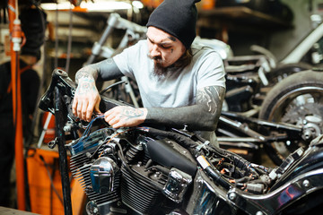 Fototapeta na wymiar Portrait of modern heavily tattooed man assembling custom motorcycle in garage in hard lighting