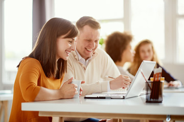 Young happy people discussing business standing at the whiteboard in modern light office, beautiful woman sitting at the table at forefront