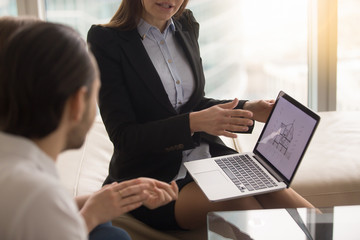 Female real estate agent offering young married couple of buyers house for sale, showing cross-sectional home plan on laptop screen, discussing house buying, considering home purchase, close up