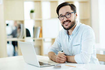 Portrait of Asian adult man smiling and looking at camera while working with laptop in sunlit office