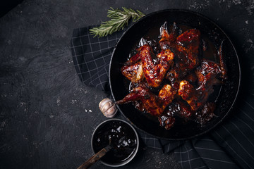 Chicken wings in thick barbecue sauce with garlic on black cast iron pan on dark rustic background.