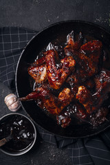 Chicken wings in thick barbecue sauce with garlic on black cast iron pan on dark rustic background.