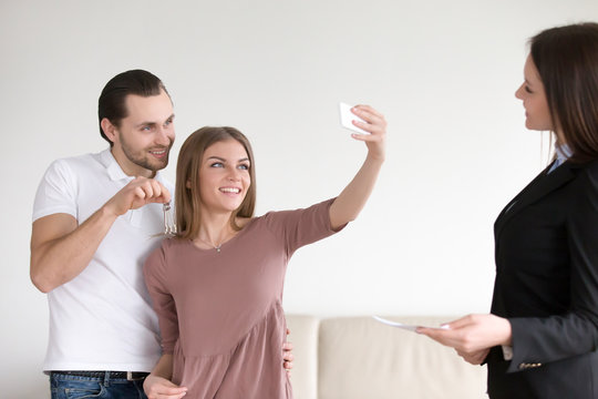 Happy apartment owners making selfie on mobile phone, showing keys to own flat, standing next to real estate agent. Young couple decided to take self-portrait photo after closing real estate deal