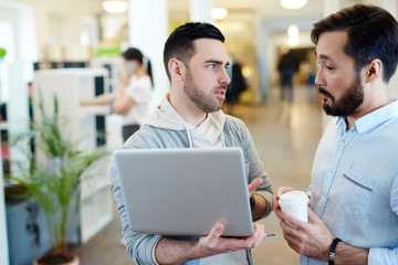 Two modern business people discussing work project standing with laptop in open office space