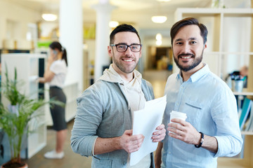 Portrait of two modern professionals looking at camera and smiling standing in open office space