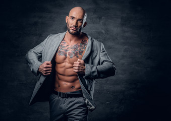 Shaved head tattooed male dressed in a grey jacket on a naked torso.