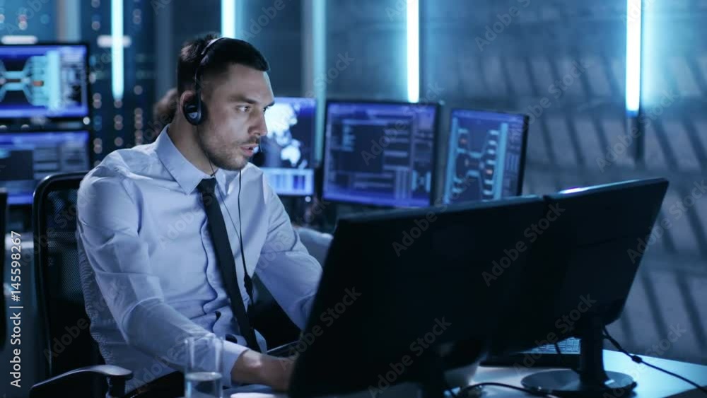 Poster in system control center technical support specialist speaks into headse while sitting at his desk b