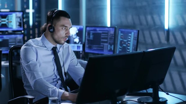 In System Control Center Technical Support Specialist Speaks into Headse while Sitting at His Desk Before Multiple Monitors. Shot on RED EPIC-W 8K Helium Cinema Camera.