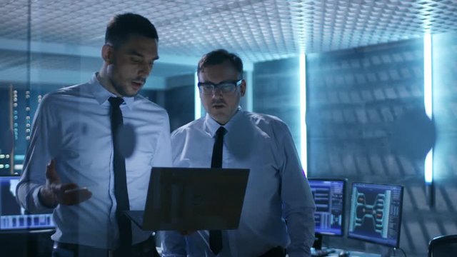 Two Security Engineers Walking In System Control Center. They Have Discussion, One of them Holds Laptop. Room is Full of Working Displays. Shot on RED EPIC-W 8K Helium Cinema Camera.