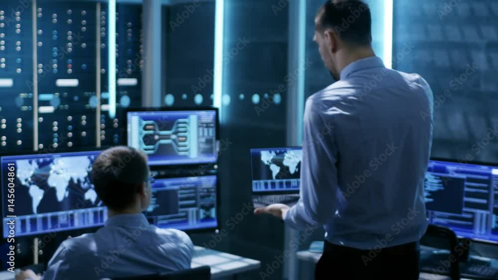 Aufkleber in system monitoring room technical engineer with laptop approaches colleague who is at his workstat