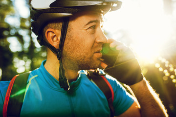 Close up portrait of young Caucasian cyclist in sportswear wearing helmet and blue t-shirt, talking on mobile phone with friend looking away, having small break while exercising outdoors in forest. 