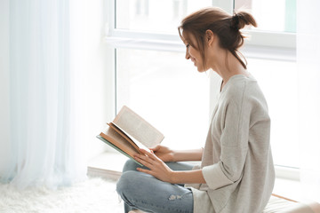 Beautiful young woman reading book near window at home - Powered by Adobe
