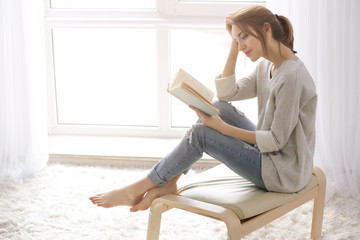 Beautiful young woman reading book near window at home