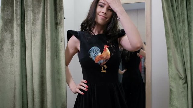 Young woman posing in dress standing in fitting room store. Spectacular brunette chooses fashionable clothes in store and cute posture in front of friend. Dressed black velvet attire with bright print