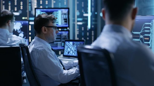 In the Monitoring Room Team of System Engineers Working at Their Workstations. Multiple Displays Show Various Important Data and Information. Possible Air Traffic/ Power Plant/ Security Room Theme. 