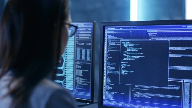 Over The Shoulder Footage of Female IT Engineer Working in Monitoring Room. She Works with Multiple Displays. Shot on RED EPIC-W 8K Helium Cinema Camera.