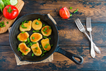 Crisp chicken nuggets with green onion in black cast iron skillet, vintage forks and fresh tomatoes and cucumbers on the cutting board. Wooden table, top view.