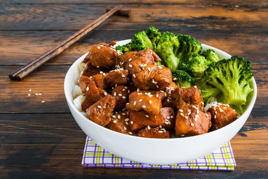 Chicken lacquered with a sweet soy teriyaki sauce in a white bowl. Garnished with rice and broccoli. Chopsticks, brown wooden table.