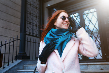 Red haired beautiful girl is walking by the street in a pink coat and blue scarf, with sunglasses.