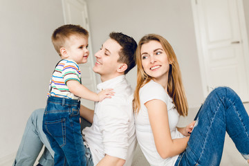 Close-up of family picture of two young parents playing with their boy child. They sit on the floor and father holds his son. They wear white t-shirt and jeans.