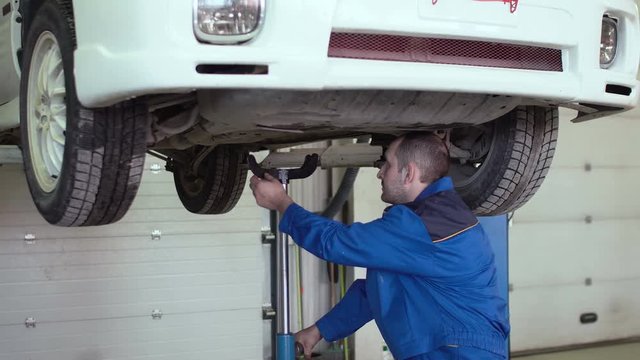 car mechanic is checking car suspension on lifted crossover car in blue overalls in car repair shop