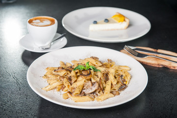 Vegeterian tasty dinner - creamy mushroom penne pasta, cup of coffee and fruit cheesecake on dark table. Close up, selective focus